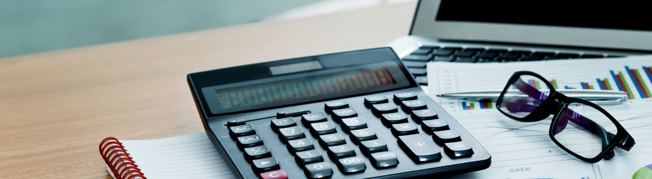 Desk with a calculator sitting on top of a notepad, next to a laptop and glasses.